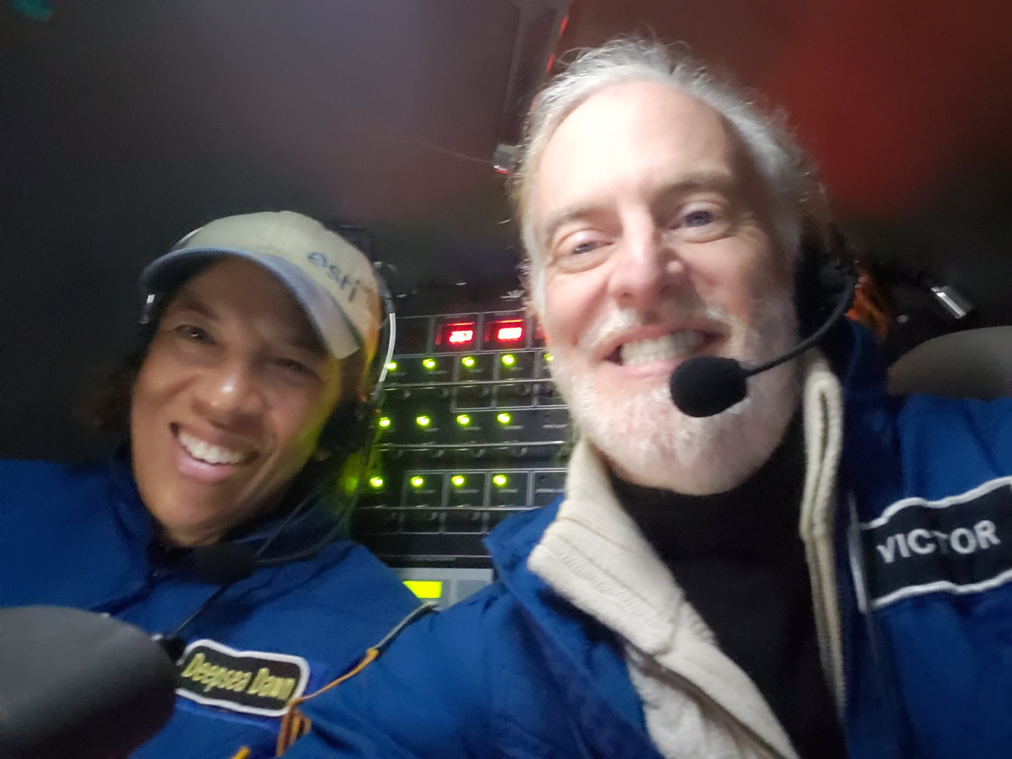 Dan and Victor smile for a selfie inside the sub. Green and red lights from the subs controls are behind them.