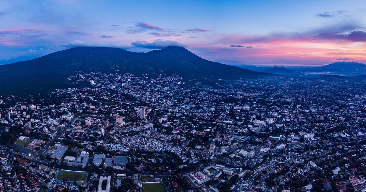 A sweeping aerial view captures a densely populated city at dusk, nestled at the base of a large mountain