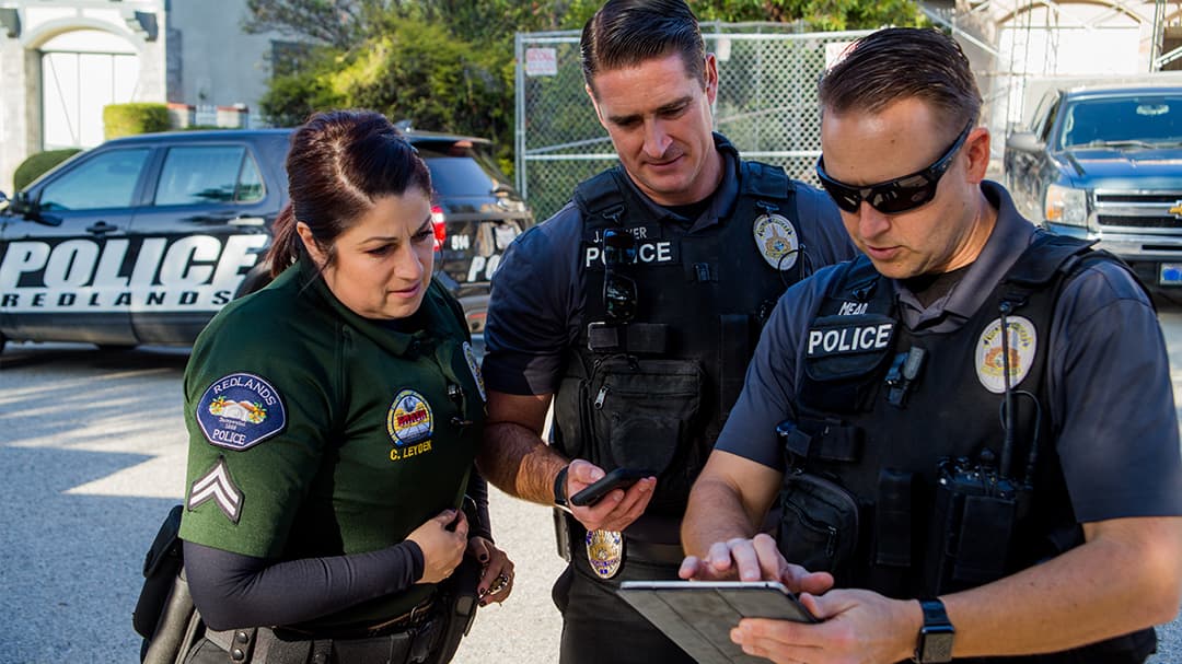 Three uniformed police officers standing in a parking lot discussing a shared tablet display