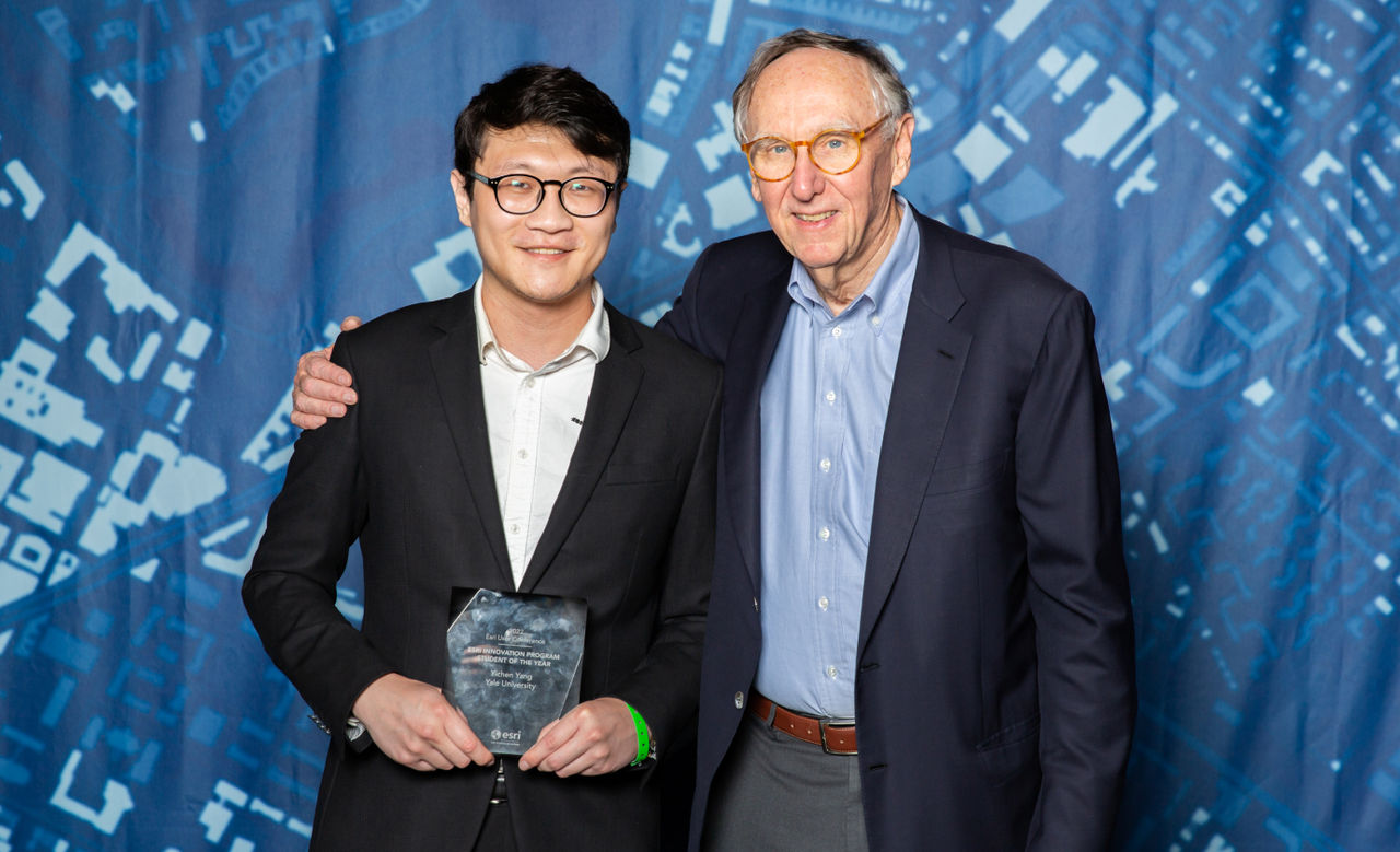 Yang holds his glass award and stands next to Dangermond stand posing for the camera