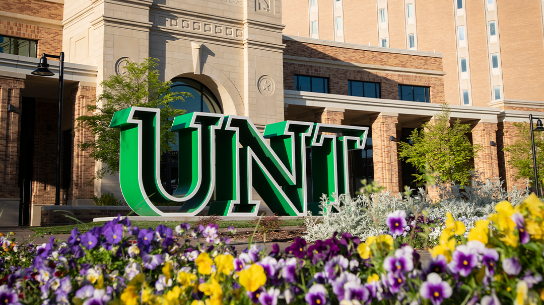 Green and white metal art sign of the university’s initials UNT in front of a campus building