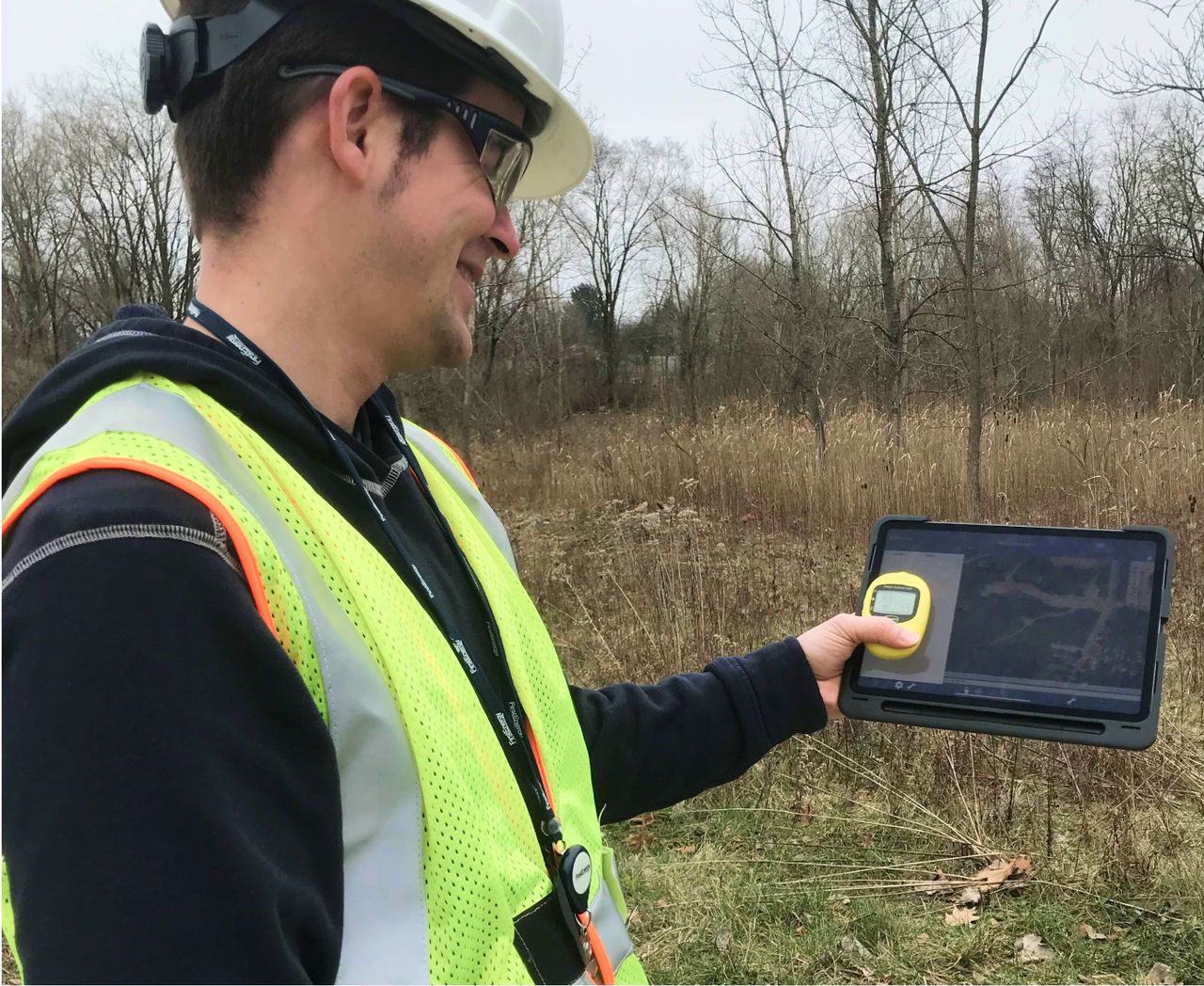 A First Energy employee wearing a yellow vest and white safety helmet, standing in a field holding an iPad