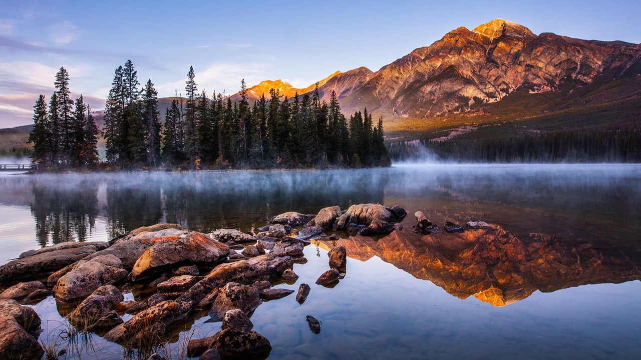 Lago alpino tranquilo que refleja árboles y montañas circundantes con rocas en primer plano