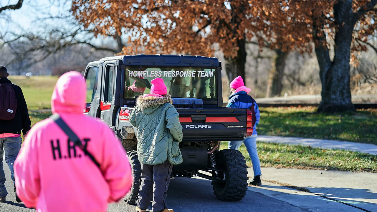 Plusieurs personnes habillées chaudement se tenant à côté du véhicule d’une équipe d’intervention pour sans-abri, à côté d’un parc ensoleillé en automne