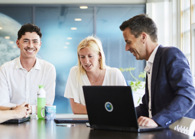 Drie collega’s werken samen aan een laptop en bespreken informatie aan een vergadertafel in een kantooromgeving.