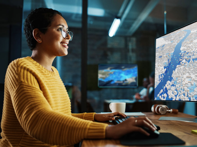 A woman in a yellow sweater uses a computer mouse and keyboard to navigate a 3D map displayed on a computer screen.