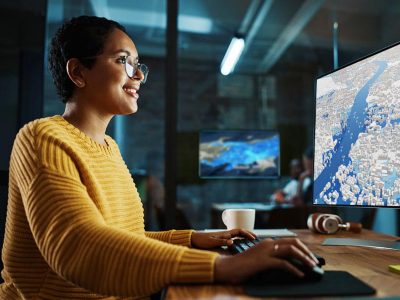 Une femme en pull jaune utilise la souris et le clavier de son ordinateur pour naviguer sur une carte 3D affichée à l’écran.