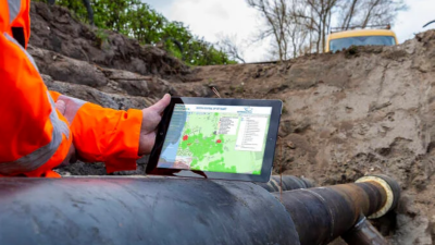 Werker in een feloranje jas houdt een tablet boven een grote ondergrondse pijpleiding in een uitgegraven sleuf. Op het scherm is een kaart te zien met gekleurde vlakken en datalagen, gebruikt voor het visualiseren en beheren van infrastructuur. 