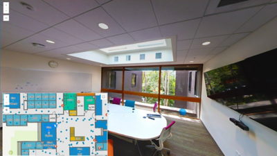 Conference room with a large white table, chairs, whiteboard, and wall-mounted monitor with an inset showing a digital floor plan.