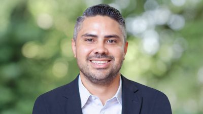 John smiling in a business suit and standing in front of green foliage in soft focus
