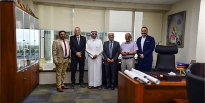 Six men pose in an office with a window in the background and a desk with rolled documents in the foreground
