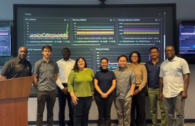 A team of nine professionals pictured in front of a large monitor displaying key performance indicators such as CPU utilization, memory utilization, and available storage capacity.
