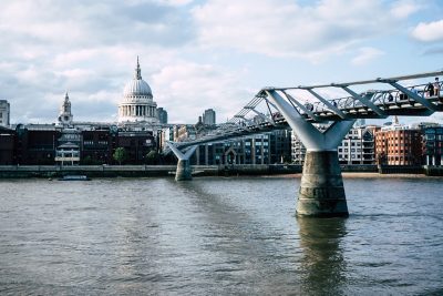 A view across the River Thames featuring the Millennium Bridge and the dome of St Paul’s Cathedral in the background under a partly cloudy sky.
