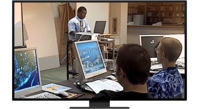 Two people sitting at desks with laptops, looking at an instructor at the front of the room