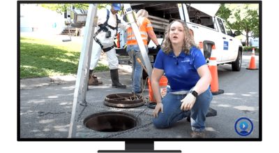 A person kneeling next to a utility maintenance hole with two workers and a utility truck in the background