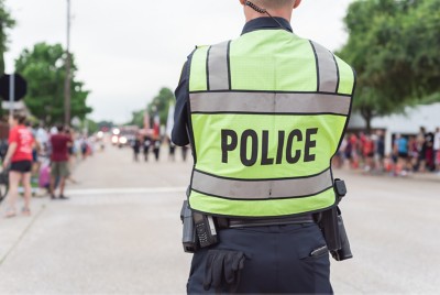 Rear view of a police officer in a reflective vest with "POLICE" printed on the back, standing in front of a crowd on a street.