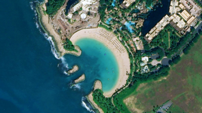 Aerial view of a beach resort with a sandy cove, turquoise water, breakwaters, buildings, and lush vegetation