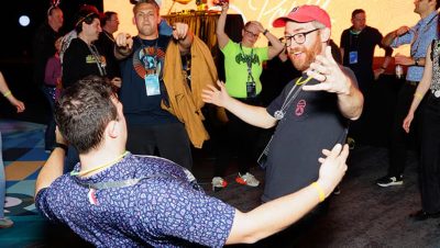A lively crowd of people dance and socialize in a packed room with a stage and vibrant lighting visible in the background