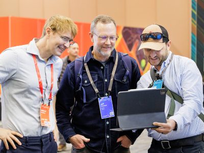 Smiling Esri expert stands with two event attendees as one holds up a tablet in an event hall