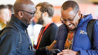 An event attendee wearing a sweatshirt and a backpack talks with another attendee in front of a colorful backdrop
