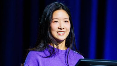 A presenter with shoulder length black hair wearing a vivid purple blouse against a dark blue background