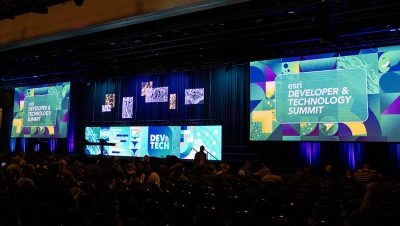 A large conference hall features a brightly lit stage with multiple wide screens displaying the Esri Developer and Technology Summit