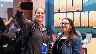 Two women smile as they take a selfie with a smartphone in front of a colorful event backdrop