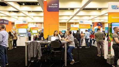 A busy technology conference expo hall features multiple booths and workstations with attendees collaborating and conversing