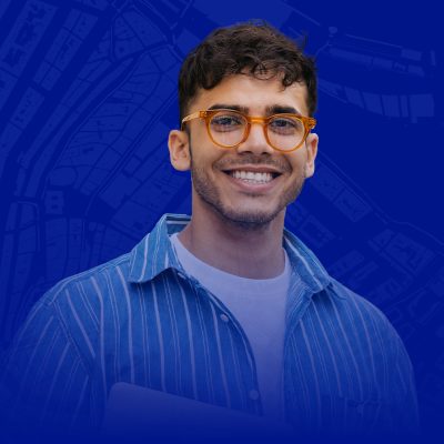 Smiling, beared person wearing glasses and a plaid shirt, with an open laptop against a map background and a stylized development symbol