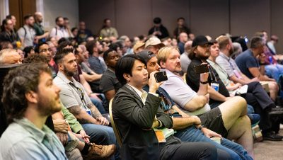 A large group of people sit closely together in a conference room, focused on a presentation out of frame
