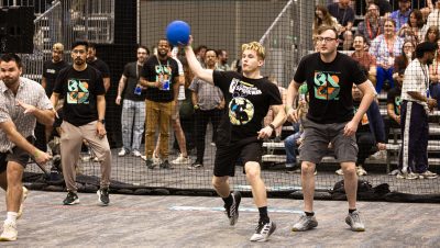 A group of adults play an indoor dodgeball game on a court surrounded by netted barriers