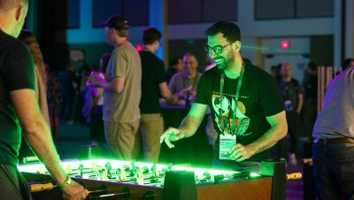 A man wearing glasses and a conference badge plays foosball on a glowing table