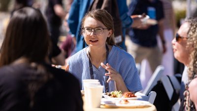 A young woman wearing glasses and a striped blue top speaks animatedly while seated at a round table outdoors