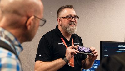 A bearded man wearing glasses and a black polo shirt presents a smartphone to another attendee in an indoor setting