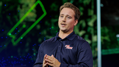 Presenter in a dark blue shirt speaking against a blurrd green map background