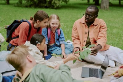Four students and a teacher sit outdoors on a picnic blanket. The teacher is showing a leafy sample to the students, who are leaning in curiously. A laptop accompanies the lesson.