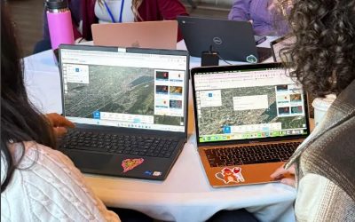 Four women sit around a table, two in the foreground and two in the background. All four women have open laptops in front of them. The women in the foreground are using their laptops to operate ArcGIS software.