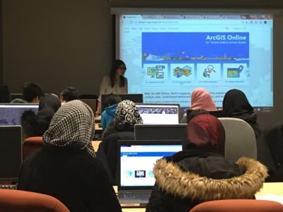 Women with laptops sit in a classroom facing the instructor at the front of the room. The instructor is teaching from a projector screen with an ArcGIS Online resource displayed on it.