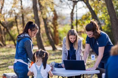 En pleine nature, deux enseignantes aux cheveux longs, munies d'un porte-nom, sont penchées sur un ordinateur portable posé sur une table pliante. De l'autre côté de la table, deux élèves, l'une préadolescente et l'autre beaucoup plus jeune, examinent avec intérêt les objets posés sur la table.
