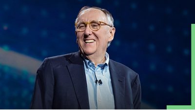 A photo of Esri founder Jack Dangermond wearing a blue shirt and blazer, smiling onstage at an Esri User Conference