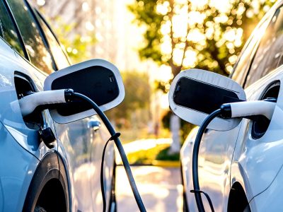 Closeup of two electric vehicles plugged into charging stations in a sunlit tree-lined parking lot