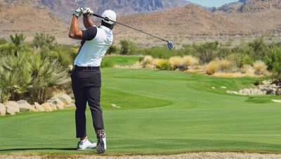 Golfer in mid-swing on a lush green golf course with desert shrubbery and a mountainous landscape in the distance