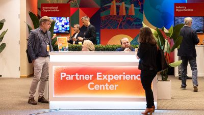 Business professionals gather around a brightly lit Partner Experience Center reception desk at a conference or trade show