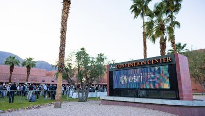 A group of attendees lines up outside the Palm Springs Convention Center near an electronic display.