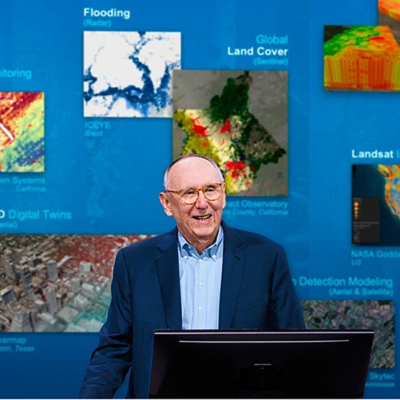 Jack Dangermond wearing a blue suit standing at a podium on stage against a backdrop of detailed digital maps
