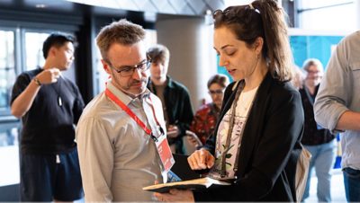 A woman shows information on a tablet to a man wearing a conference badge and lanyard in a bright indoor venue 