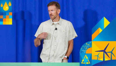 Presenter wearing a beige button up shirt and a lapel microphone in front of a blue curtain