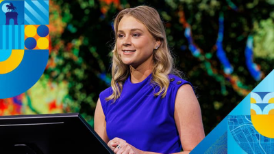 Presenter wearing a royal blue blouse speaking from a podium with a large screen behind displaying a detailed map