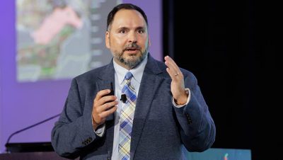 Presenter in a suit and tie gestures with both hands while speaking on stage