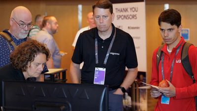 A group of event attendees looks on as an exhibitor works on a laptop in the Expo area